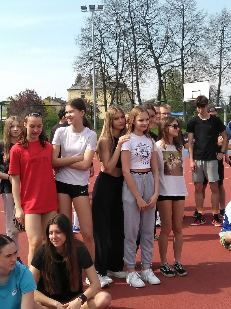 Grupa około 10 dziewcząt siedząca i stojąca na boisku. W tle kilku chłopaków