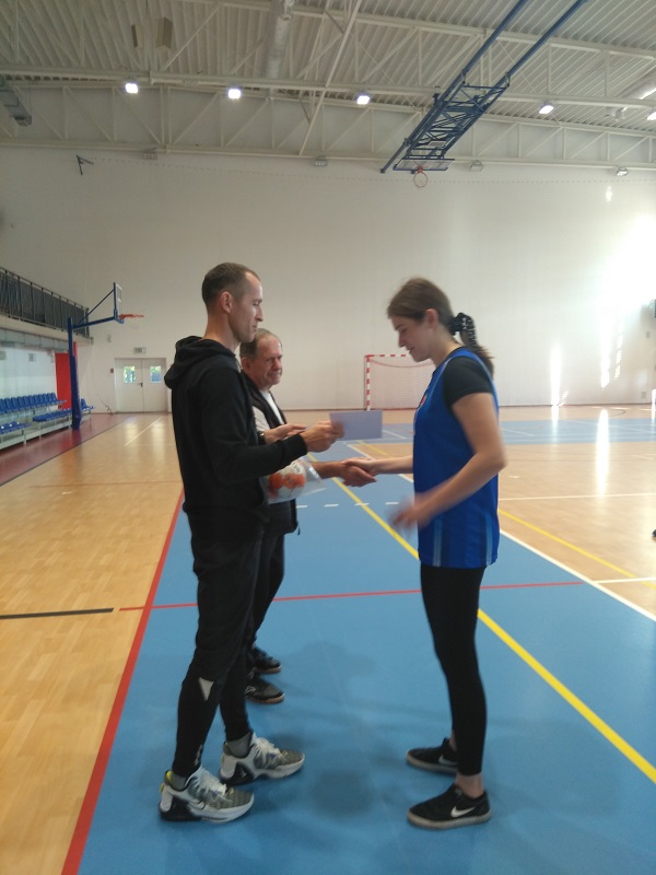 Uczennica na hali sportowej odbiera dyplom z rąk dwóch gratulujących jej mężczyzn.